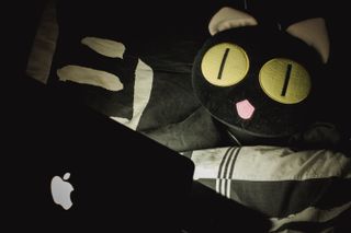 A plush cat on a bed watching the screen of a laptop computer
