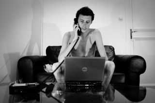 A nude man on a sofa with a phone on his ear and a computer in front of him